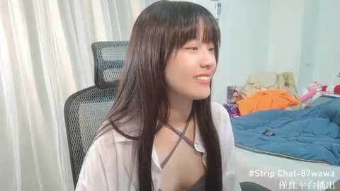 Media: A video of a smiling Asian woman with long black hair, wearing a white shirt, in a messy bedroom.