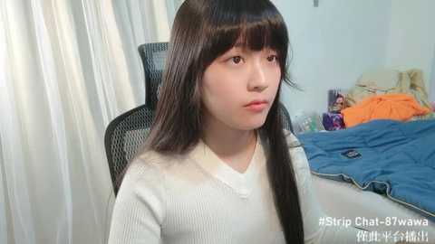 Media: Video of a young Asian girl with long black hair, wearing a white sweater, sitting on a black chair in a messy bedroom with blue bedding and scattered clothes.