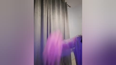 Media: Video of a person wearing a purple shirt and gray pants, standing in front of a gray curtain in a dimly lit room.