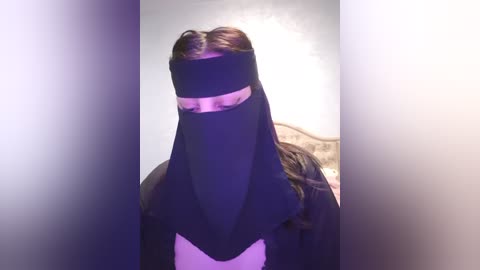 Media: A video of a person wearing a black niqab with a pink bandana over the face, set against a soft, blurred background with a hint of a bed and wall.