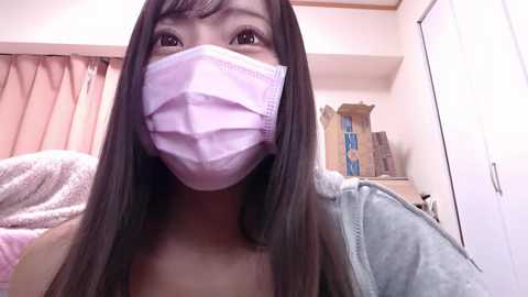 Media: A video of an Asian woman with long black hair, wearing a pink surgical mask, light blue jacket, and a beige towel. Background shows a bathroom with beige curtains and white walls.