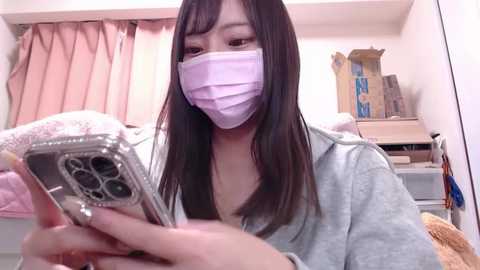 Media: A video of an East Asian woman with long black hair and a light complexion, wearing a pink surgical mask and gray hoodie, examining a smartphone in a cluttered bedroom with pink curtains, cardboard boxes, and a stuffed teddy bear.