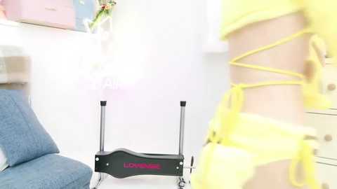 Media: Video of a person wearing a yellow, lace-up bodysuit, standing in a brightly lit room with a blue chair, white wall, and a black mobility aid with \"Lift\" branding.