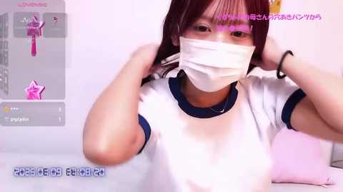 Video of an East Asian woman with light skin and straight, shoulder-length brown hair, wearing a white T-shirt, blue trim, and a white mask, adjusting her hair. Background shows a pink character and a smartphone screen with Japanese text.