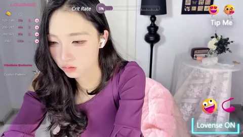 Media: A video of an Asian woman with long, wavy black hair, wearing a purple sweater, sitting in a cozy room with a floral-patterned table, a black lamp, and a framed picture.