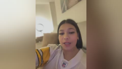 Media: A video of a young Asian woman with straight black hair, wearing a white hockey jersey with yellow stripes, smiling and holding a camera. Background features a beige couch and a dimly lit room.