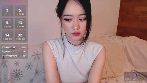 Media: Video of a young East Asian woman with pale skin and long black hair, wearing a sleeveless white top, sitting on a bed with floral sheets in a cozy, warmly lit bedroom.