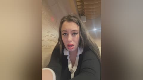 Media: Video of a young woman with long brown hair, wearing a black cardigan over a white blouse, with a distressed expression, partially obscured by a glass partition.