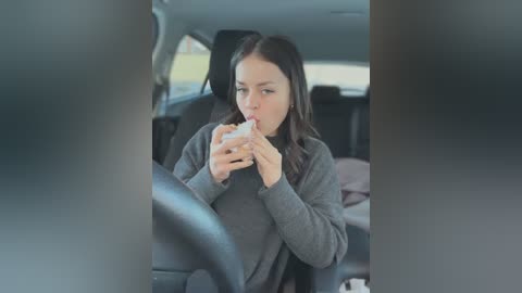 Media: Video of a young woman with long dark hair, wearing a gray sweater, eating a sandwich while driving a car. The car's interior is visible with a dark headrest and window.