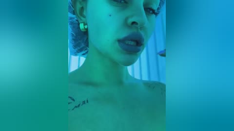 Media: Video of a young woman with light skin, curly hair, and a tattoo on her shoulder, in a dimly lit room with blue-green lighting.