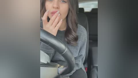 Media: Video of a young woman with long brown hair, wearing a gray sweater, sitting in a car, hand to her mouth, steering wheel in focus, blurred background.