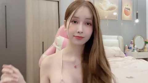 Media: A video of a young, fair-skinned East Asian woman with long brown hair, wearing a pink gaming headset, in a modern, minimalist bedroom with white furniture, peach-themed artwork, and a neatly made bed.