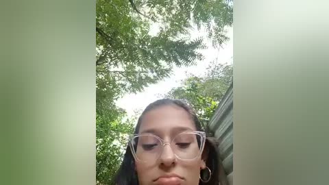 Media: A video of a young woman with light brown skin and medium-length black hair, wearing large, clear-rimmed glasses, captured from a low angle. She stands in a lush, green forest with tall trees and bright sunlight filtering through the foliage.