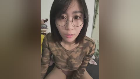 Media: Video of an East Asian woman with short black hair, wearing glasses, and a camouflage-patterned top, sitting indoors with a blurred background.