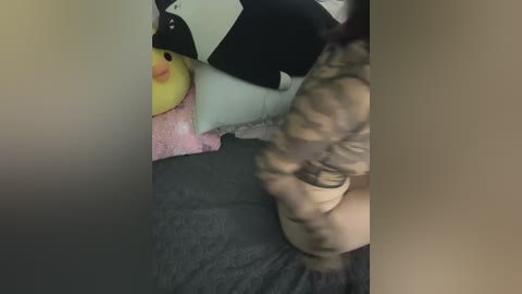 Media: Video of a partially visible person with curly hair, wearing a camouflage-patterned shirt, kneeling on a grey bedspread. A yellow duck toy and a white pillow are in the background.