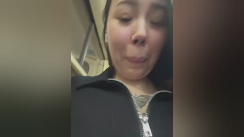 Media: Video of a young Asian woman with light skin, wearing a black jacket and a silver necklace, making a silly face in a dimly lit room.