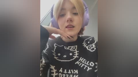 Media: Video of a young woman with short blonde hair, wearing large purple headphones, a black sweater with white Hello Kitty patterns, and black nail polish, touching her chin, indoors.