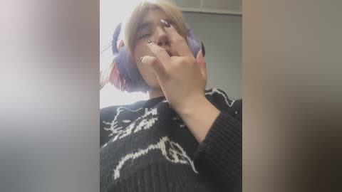 Media: Video of a person with light skin and shoulder-length blonde hair wearing large, purple headphones, covering their eyes. They are dressed in a black, knitted sweater with white bear patterns.