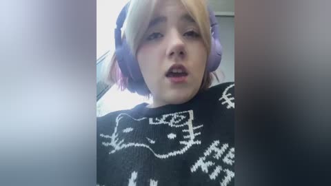 Media: A video of a young Asian woman with light skin and short, blonde hair wearing large purple headphones and a black sweater with white designs. She has a neutral expression, mouth slightly open.