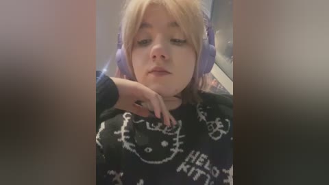 Media: A video of a young person with light skin and blonde hair, wearing purple headphones, a black sweater with white \"Hello Kitty\" designs, and a contemplative expression.