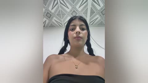 Media: Video of a young, light-skinned Latina woman with long black hair in braids, wearing a strapless black top, against a geometric-patterned white ceiling. She has a neutral expression.