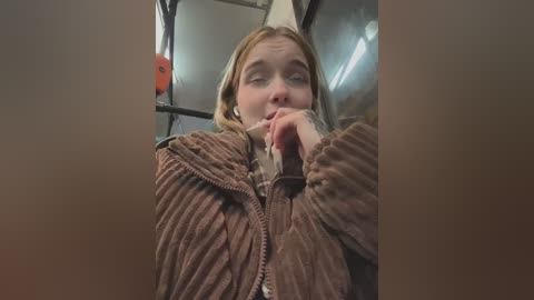 Media: Video of a young blonde woman with light skin, wearing a brown ribbed sweater, resting her chin on her hand, looking contemplative inside a dimly lit, metallic subway car.