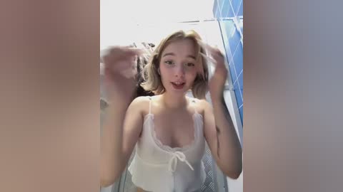 Media: Video of a young, fair-skinned woman with blonde hair, wearing a white lace lingerie top, standing in a tiled bathroom.