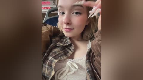 Media: A video of a young woman with light skin, blonde hair, and blue eyes, wearing a plaid shirt and white lace top, sitting on a bus. She's holding a phone, smiling, and looking at the camera.