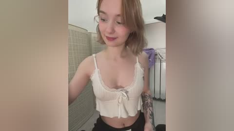 Media: Video of a young woman with shoulder-length blonde hair, fair skin, and a slender build. She wears a white lace-trimmed crop top, revealing a small tattoo on her right arm. Background features a beige wicker screen and laundry room with a metal drying rack.