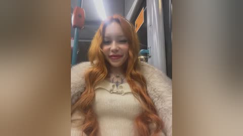 Media: A video of a smiling, light-skinned woman with long, wavy orange hair, wearing a cream sweater and a fur-lined jacket, standing in a subway car with metal rails and a red strap hanging in the background.