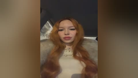 Media: Video of a young woman with long, wavy auburn hair, fair skin, and a petite physique. She wears a white top and sits on a light-colored blanket in a dimly lit room.