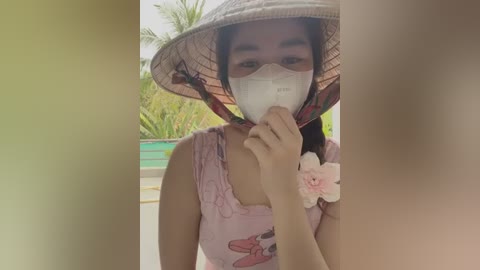 Media: Video of a woman in a pink floral dress and conical hat, wearing a face mask, standing in a garden with palm trees.