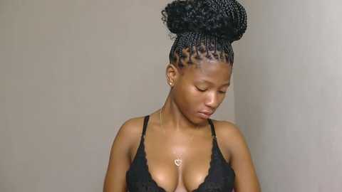 Video of a dark-skinned woman with braided hair, wearing a black lace bra, looking down with a contemplative expression against a plain, beige background.