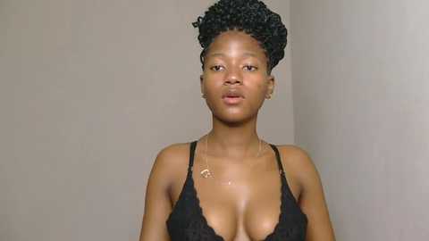 Video of a young Black woman with a slender build, medium brown skin, and natural hair styled in tight curls. She wears a black lace bralette, revealing her medium-sized breasts, and small gold hoop earrings. The background is a plain, light beige wall.