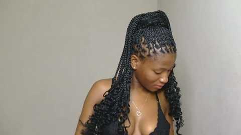 Media: Video of a young African-American woman with medium-dark skin, wearing black braids and a black halter top, standing against a plain beige wall. She has a calm expression and small earrings.