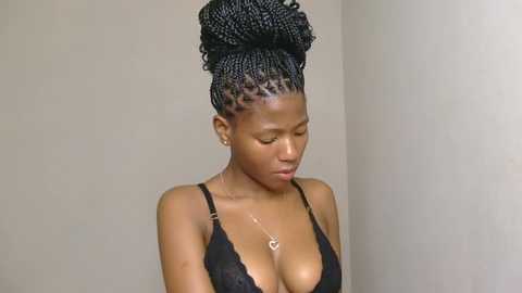 Video of a young Black woman with a medium-dark complexion, wearing a black lace bra, her hair styled in a large, intricate updo. She has a slender build and medium-sized breasts. The background is plain, off-white.