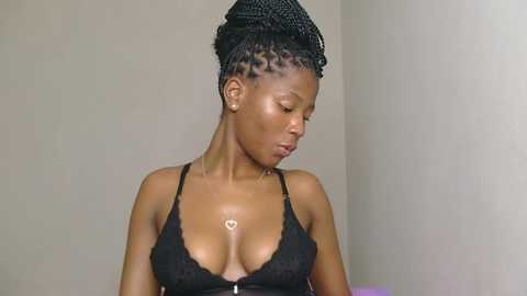 Video of a slender, dark-skinned woman with medium-sized breasts, wearing a black lace bra, her hair styled in tight braids. She has a serene expression, set against a plain beige wall.