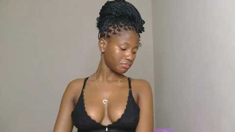 Video of a young Black woman with a medium-dark skin tone and short, styled braids. She wears a black lace bralette that accentuates her medium-sized breasts. The background is a plain, light-colored wall, creating a minimalist, uncluttered setting.