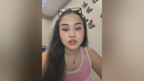 Media: Video of an East Asian woman with long black hair, wearing a pink and white striped tank top, standing in a room with butterfly stickers on the wall.