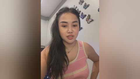 Media: Video of a young Asian woman with long black hair, wearing a pink and white striped tank top, standing in a brightly lit room adorned with butterfly wall art.