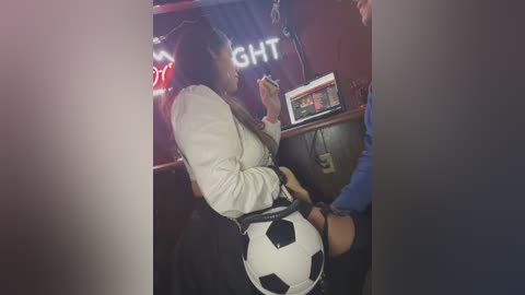 Media: Video of a woman in a white shirt and black skirt seated in a dimly lit bar, holding a soccer ball and a drink, with a man in a blue shirt standing nearby.