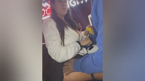 Media: Video of a woman in a white sweater being held from behind by a man in a blue hoodie, in a dimly lit bar with neon signs.