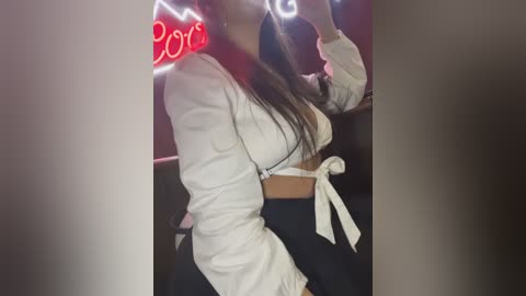 Media: A video of a woman taking a selfie indoors, wearing a white cropped top and black pants, with a red neon \"2019\" sign in the background.