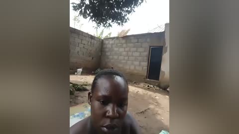 Media: Video of a young African boy with short hair, standing shirtless in a sunlit, dirt courtyard with a concrete wall, small blue door, and a small white dog in the background.