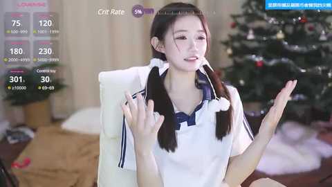 Media: A video of a young Asian woman with long dark hair in pigtails, wearing a white blouse with a blue collar, seated in front of a Christmas tree in a cozy living room.