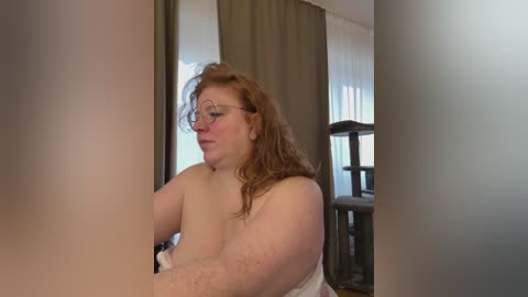 Media: Video of a plus-size, fair-skinned, red-haired woman with glasses, wearing only a towel, sitting in a dimly lit room with a dark wooden chair and beige curtains in the background.