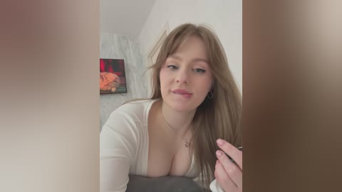 Media: A video of a young, fair-skinned woman with light brown hair and a cleavage-revealing white top, smiling while holding a phone.