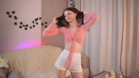 Media: Video of a slim, light-skinned woman with shoulder-length brown hair, wearing a pink crop top and white shorts, posing in a cozy living room with pastel walls, curtains, and a plush sofa.