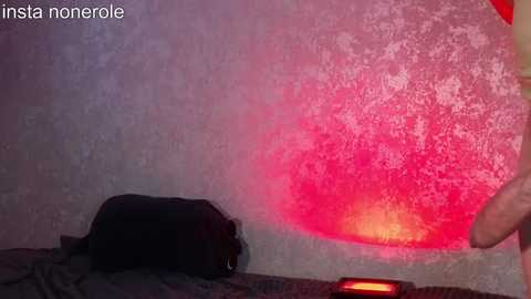 Media: Video of a dimly lit room with a black cat sleeping on a dark bedspread, a red and orange light glowing on a textured, stucco wall, and a hand holding a phone visible on the right.