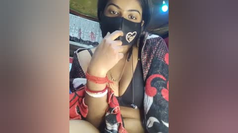 Media: Video of a woman with medium skin tone, dark hair, wearing a black mask, red heart necklace, and a black and red patterned robe. She's bound with red rope, in a dimly lit room.